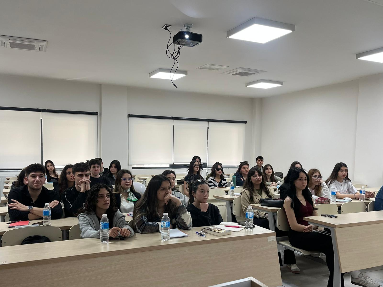 Bu fotoğraf ise sınıfın orta sıralarından bir açı sunuyor ve öğrencilerin söyleşiye olan ilgisini yansıtıyor. Öğrencilerin büyük çoğunluğunun yüzleri sahneye/ekrana dönük; kimileri dikkatle dinliyor, kimileri ise o anın bir parçası olarak objektife yansıyor. Genel atmosfer, oldukça ciddi ve akademik bir eğitim ortamını başarıyla yansıtıyor.